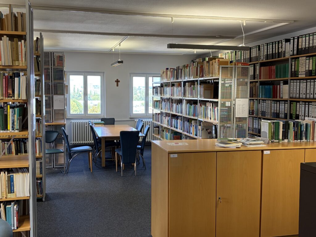 Lesesaal der CIBEDO-Bibliothek mit Regalen, Tisch und Kreuz an der Wand
