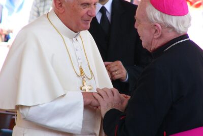 Papst Benedikt XVI. reicht einem Bischof die Hände, im Hintergrund Gläubige